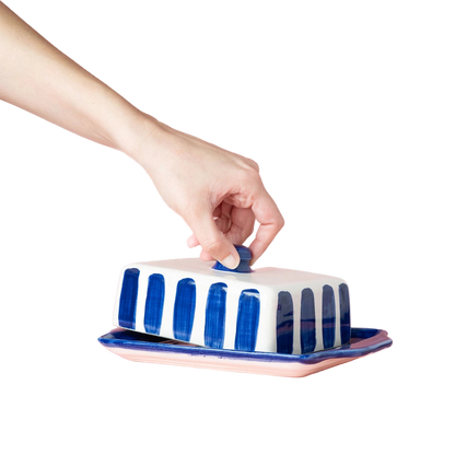 Hand closing a blue and white striped butter dish on a pink background